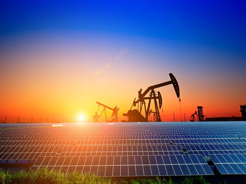 Solar panels in the foreground with oil pump jacks in the background illustrating energy transition from fossil fuels to renewable energy during sunset
